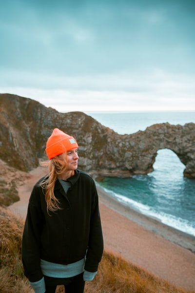 白天，一个身穿黑色夹克、头戴橘色针织帽的女人站在沙滩上